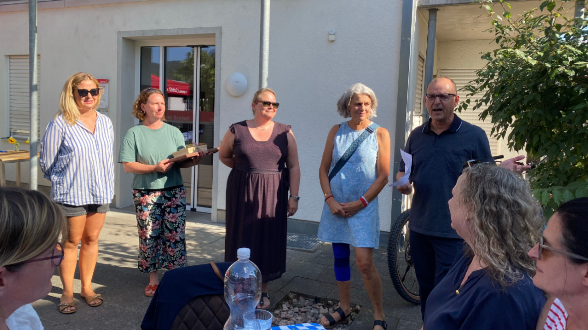 5 Personen stehen im Halbkreis und sprechen zu einer Gruppe Menschen. 